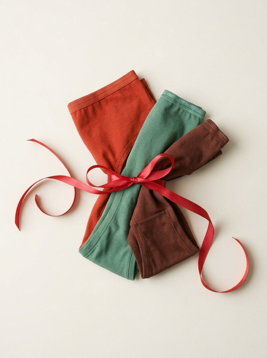 Three pairs of folded underwear rust red, sage green, and brown, tied together with a red ribbon on a light cream background.