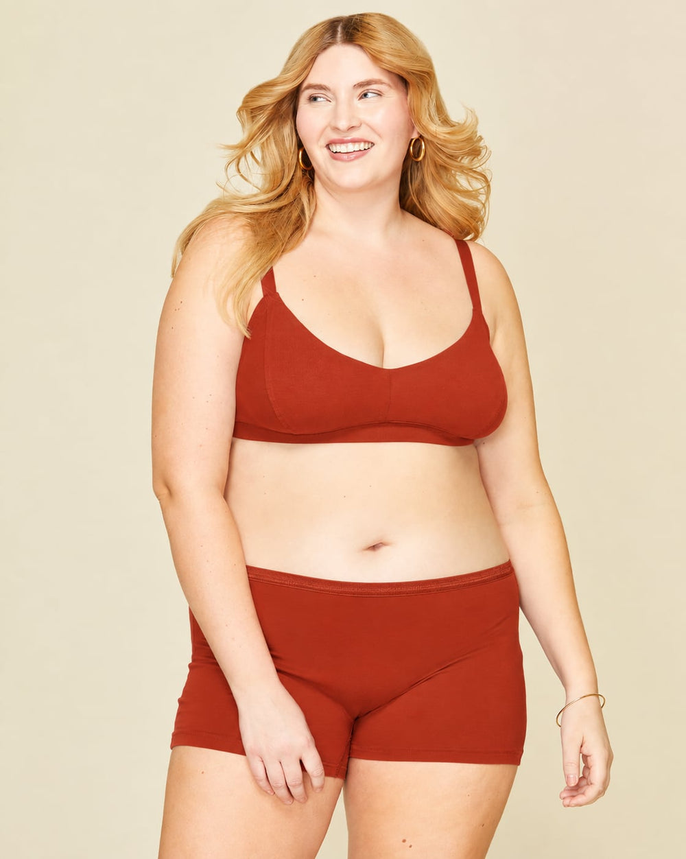 A woman modeling mid rise shorties in solid terracotta orange color, standing against a plain beige background.