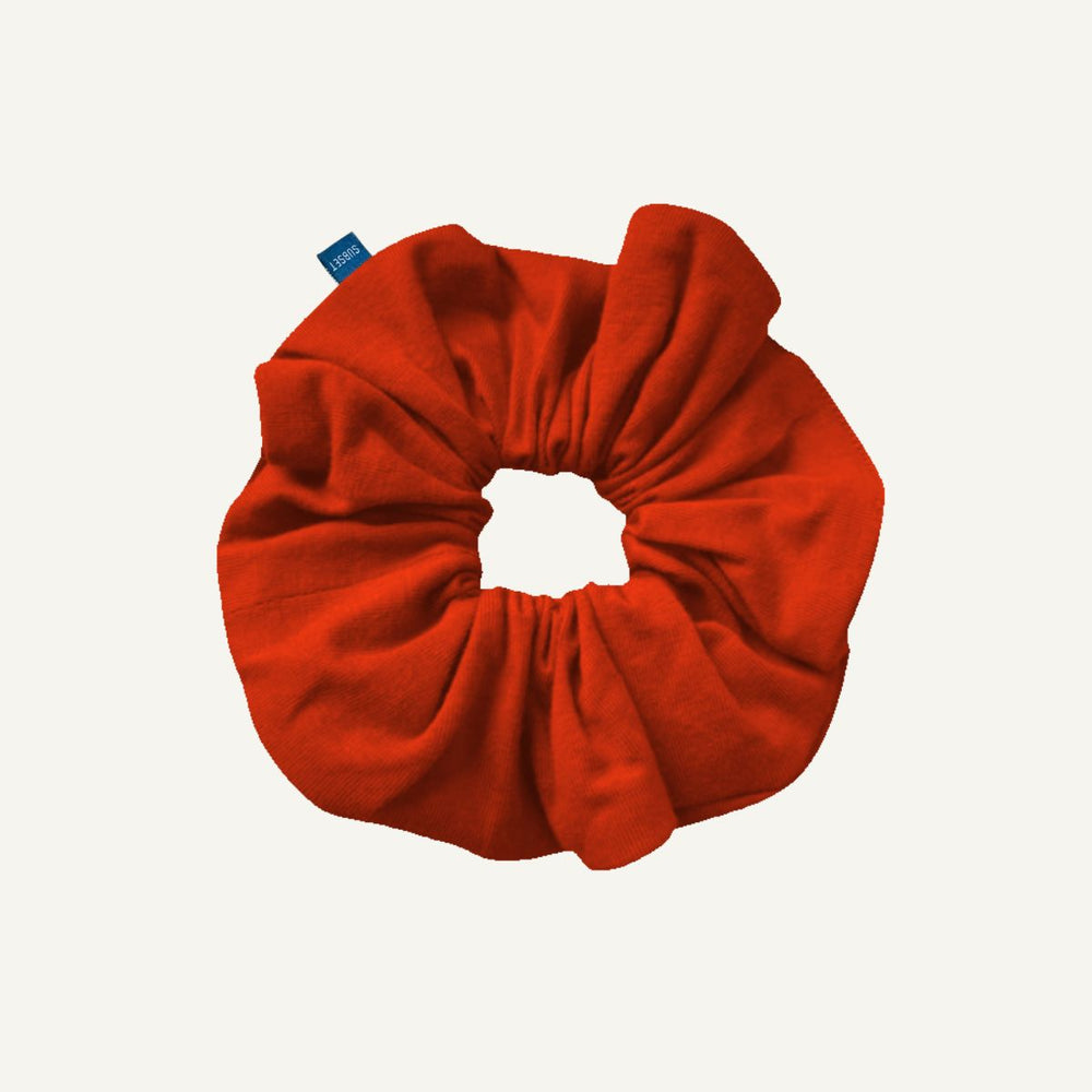 A terracotta orange fabric scrunchie with a small blue brand tag, shown on a plain off-white background.